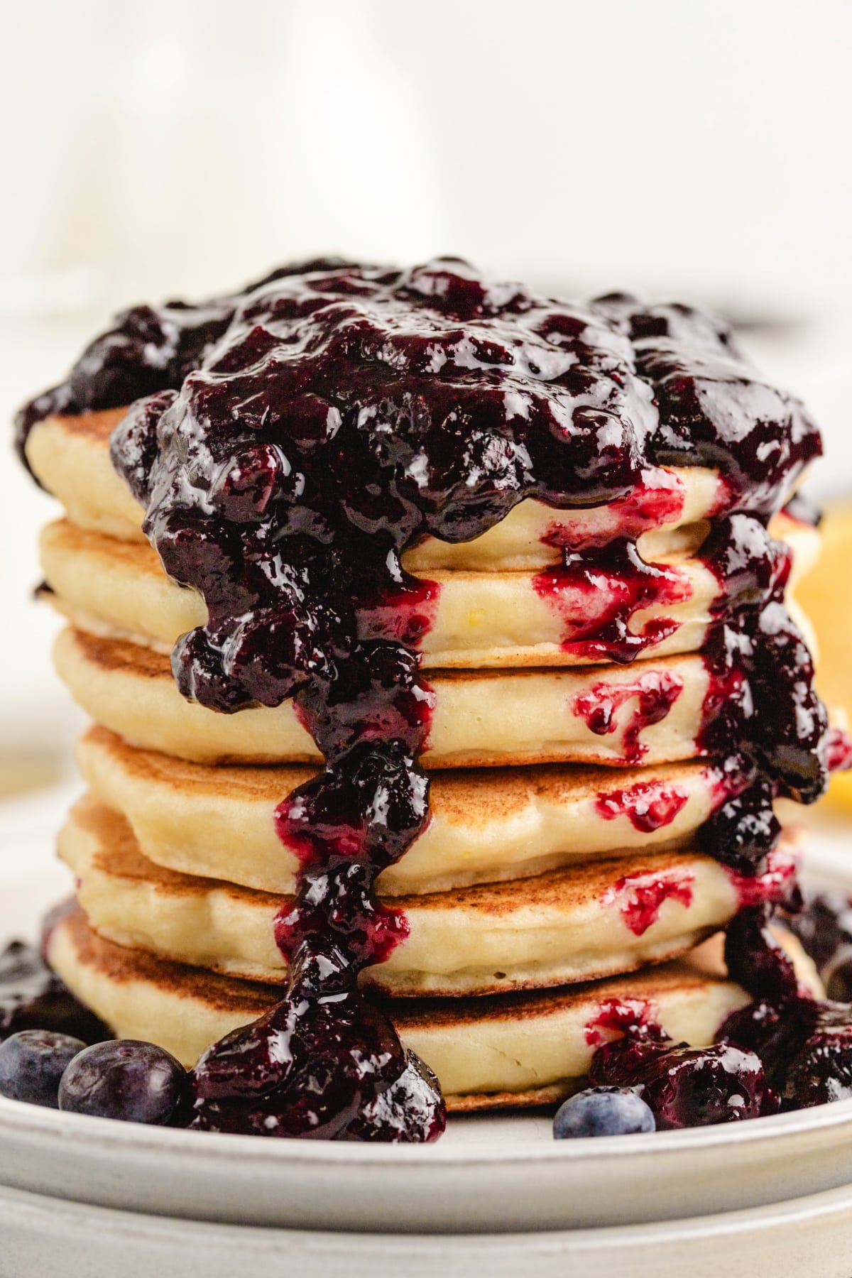 A tall stack of golden brown lemon ricotta pancakes with blueberry sauce cascading down the sides on a white plate.