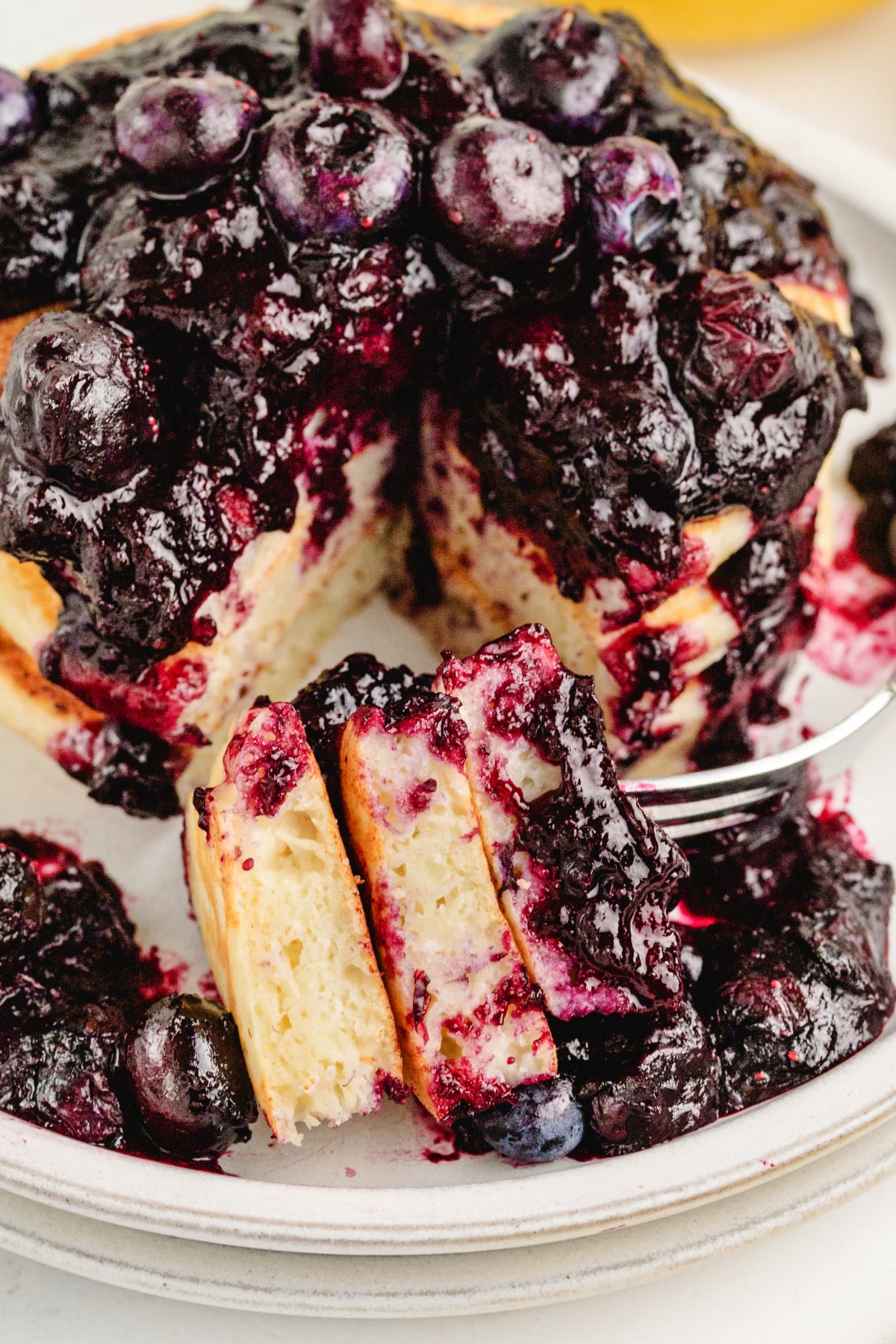 A fork holding a bite of lemon ricotta pancake covered in blueberry sauce, with the full stack visible in the background. 