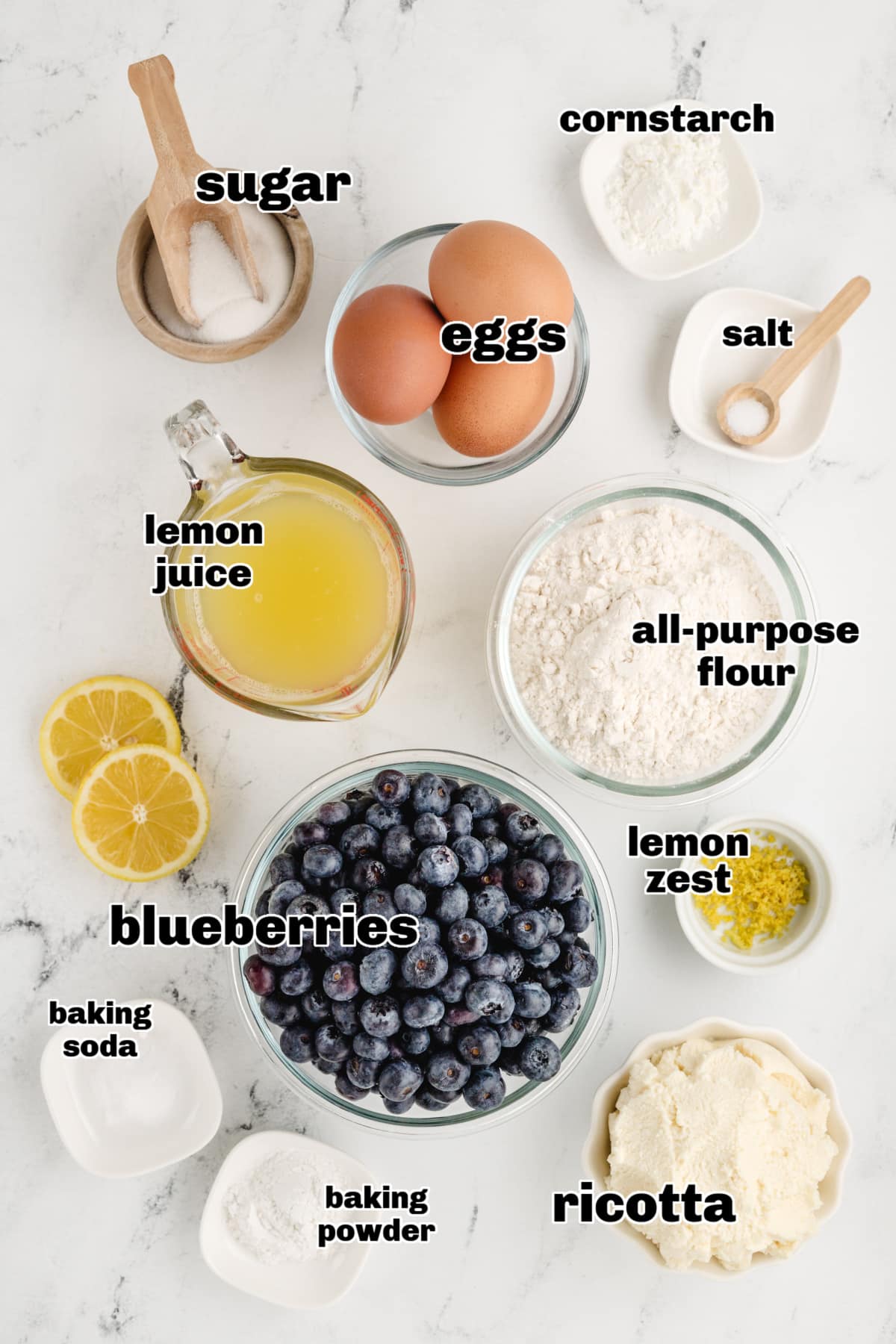 All ingredients for pancakes and sauce neatly arranged on a countertop, including flour, ricotta, eggs, lemon, and blueberries.