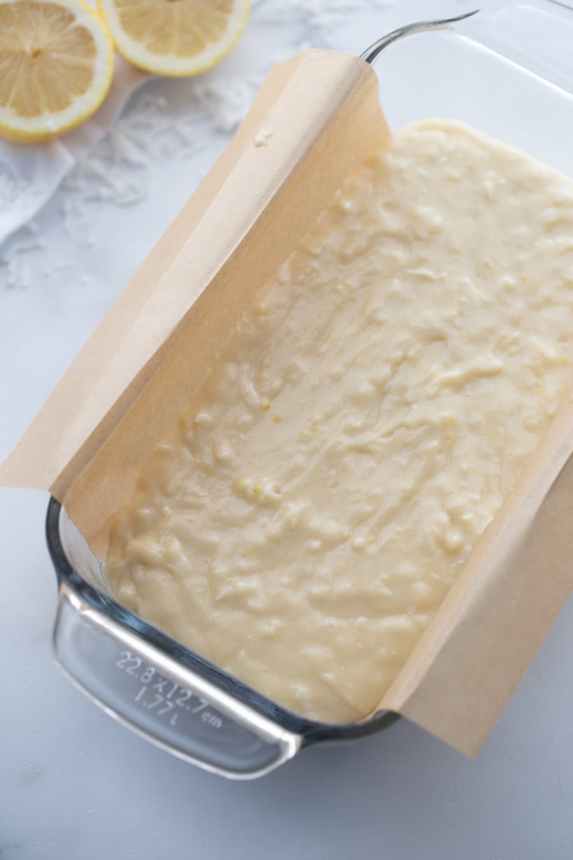 Lemon loaf batter in a baking pan with parchment paper overhang, ready for baking.