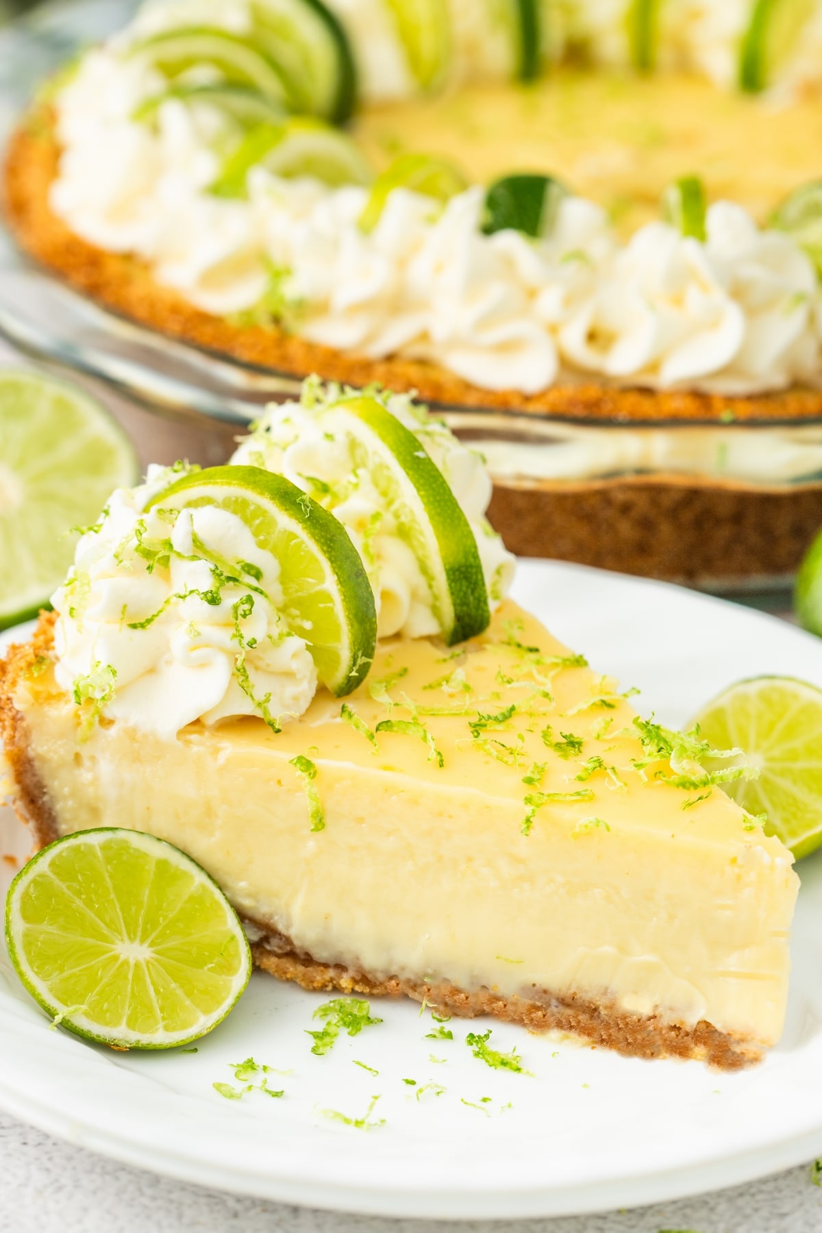 Plated slice of key lime pie with whole pie behind it Slice of key lime pie on a plate with the full pie in the background