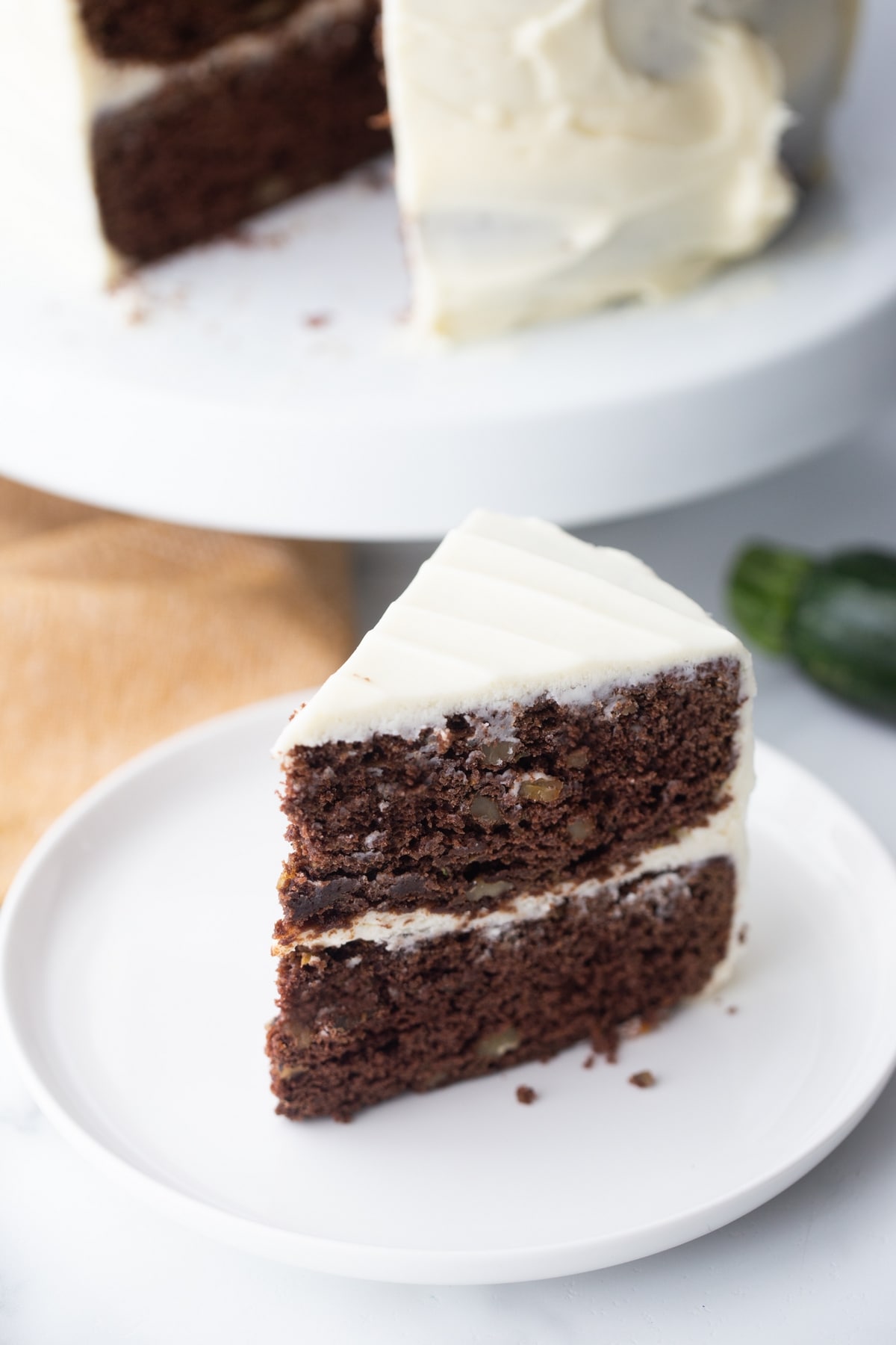 Rich slice of chocolate zucchini cake with cream cheese frosting served on a white plate, perfect example of a moist zucchini chocolate cake recipe.