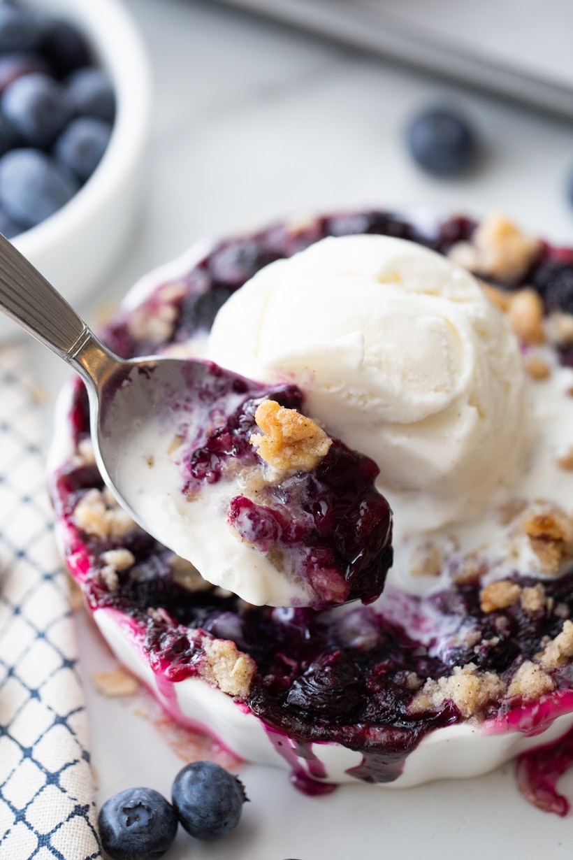 blueberry crisp with ice cream and bite taken Vanilla ice cream-topped blueberry crisp with a spoon taking a bite