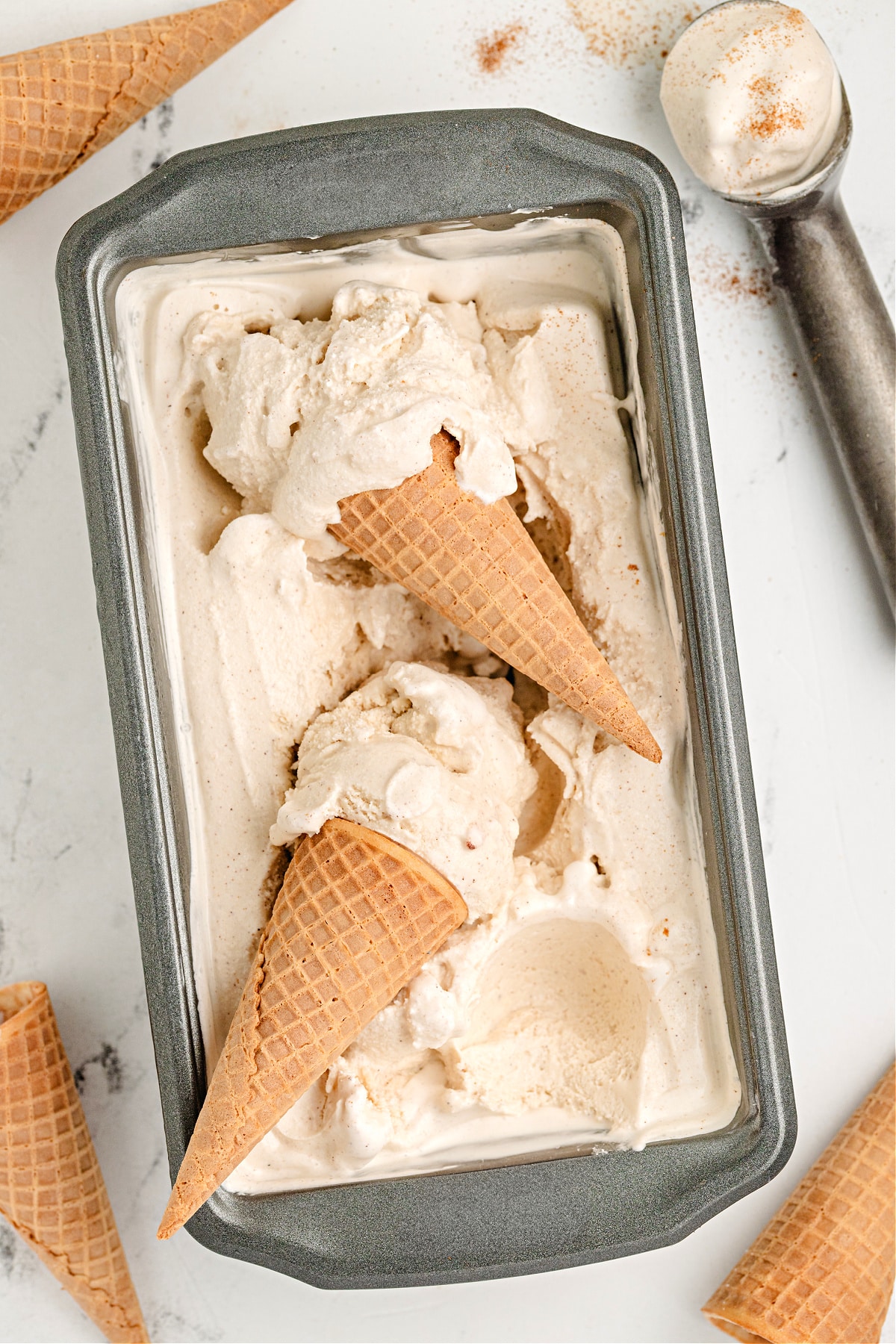 Loaf pan of churned snickerdoodle ice cream with two cones resting on top.