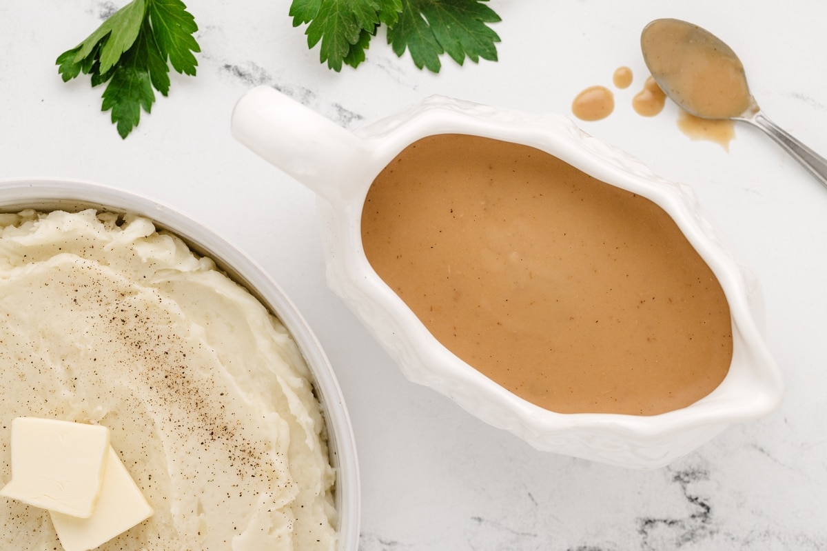 Make Ahead Turkey Gravy in White Gravy Boat Creamy make ahead turkey gravy for Noble Pig shown in a white gravy boat beside a bowl of mashed potatoes. Smooth, golden brown, and ready for Thanksgiving serving.