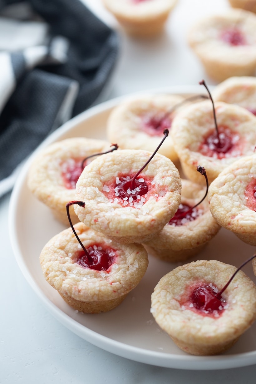 Long stemmed maraschino cherries burst right out the top of these pillowy-soft and bite-sized pastry pouch cookies. They are the perfect addition to any dessert table. cherry tassie