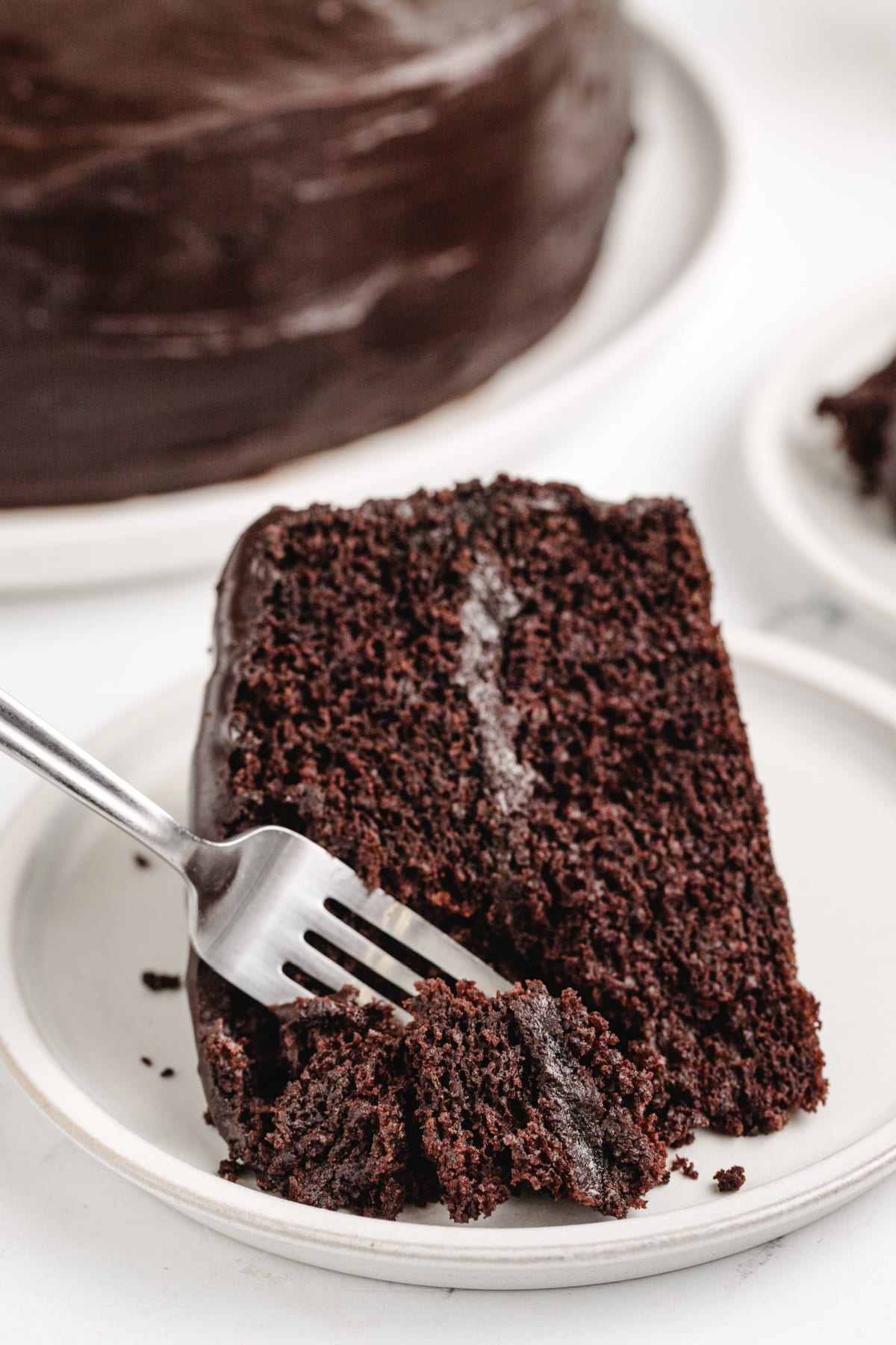 A fork cutting into a slice of chocolate layer cake, highlighting the soft texture and thick chocolate icing.
