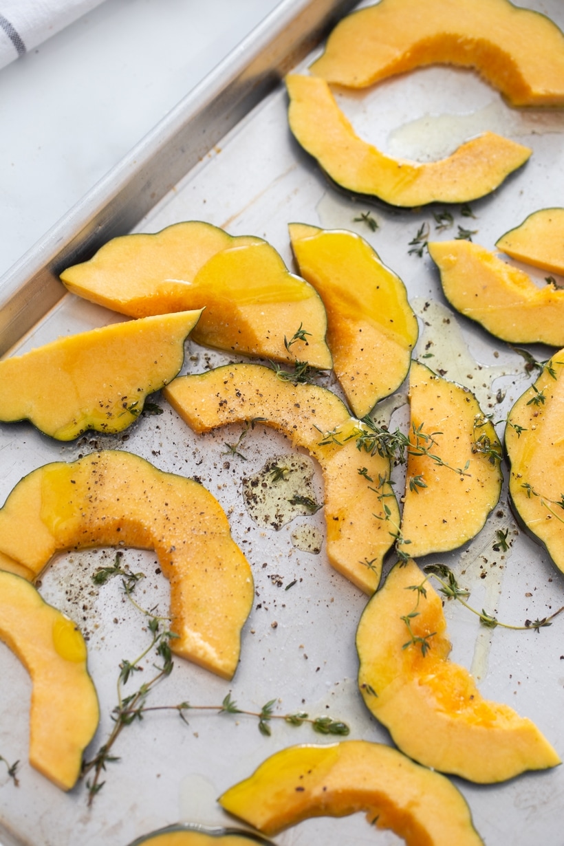 raw acorn squash slices on a baking sheet with olive oil, thyme, salt and pepper for roasting