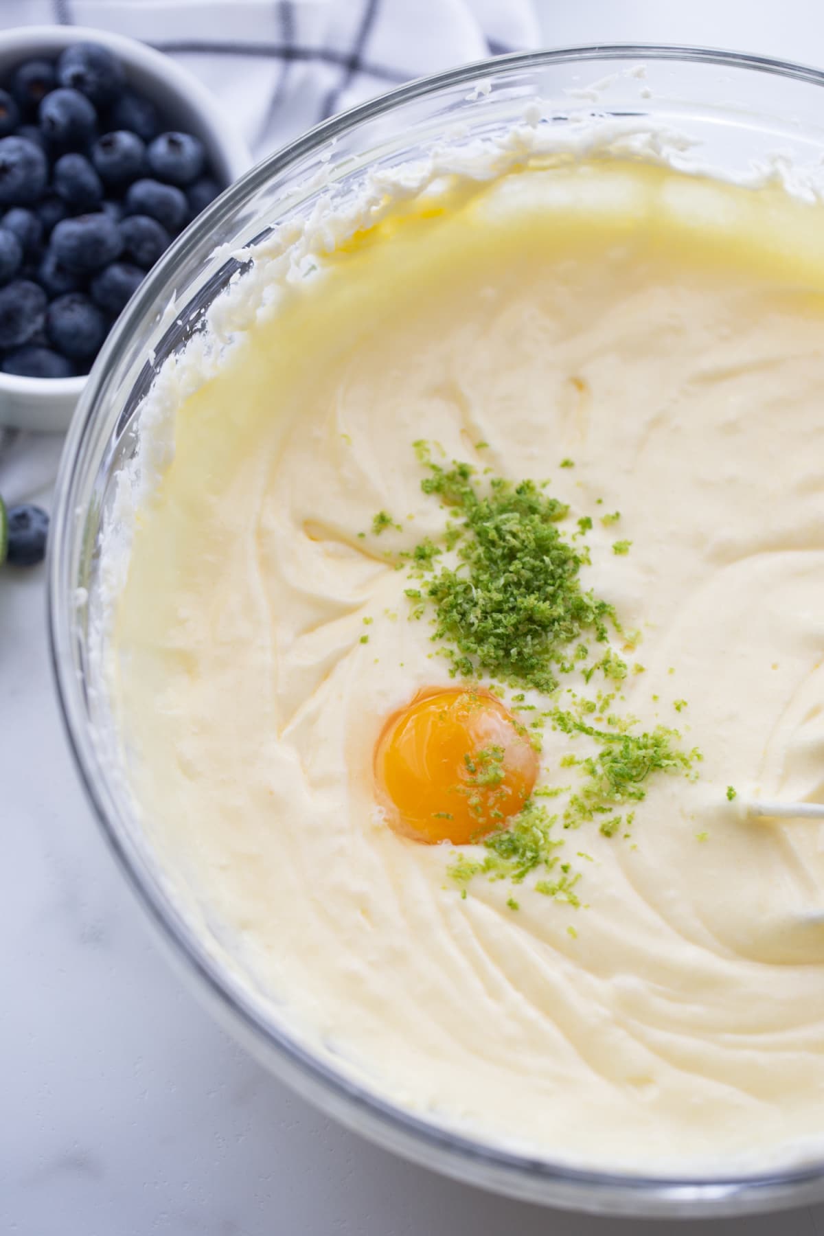 Mixing bowl filled with pale batter, with a full egg yolk and fresh lime zest sitting on top before being mixed in.
