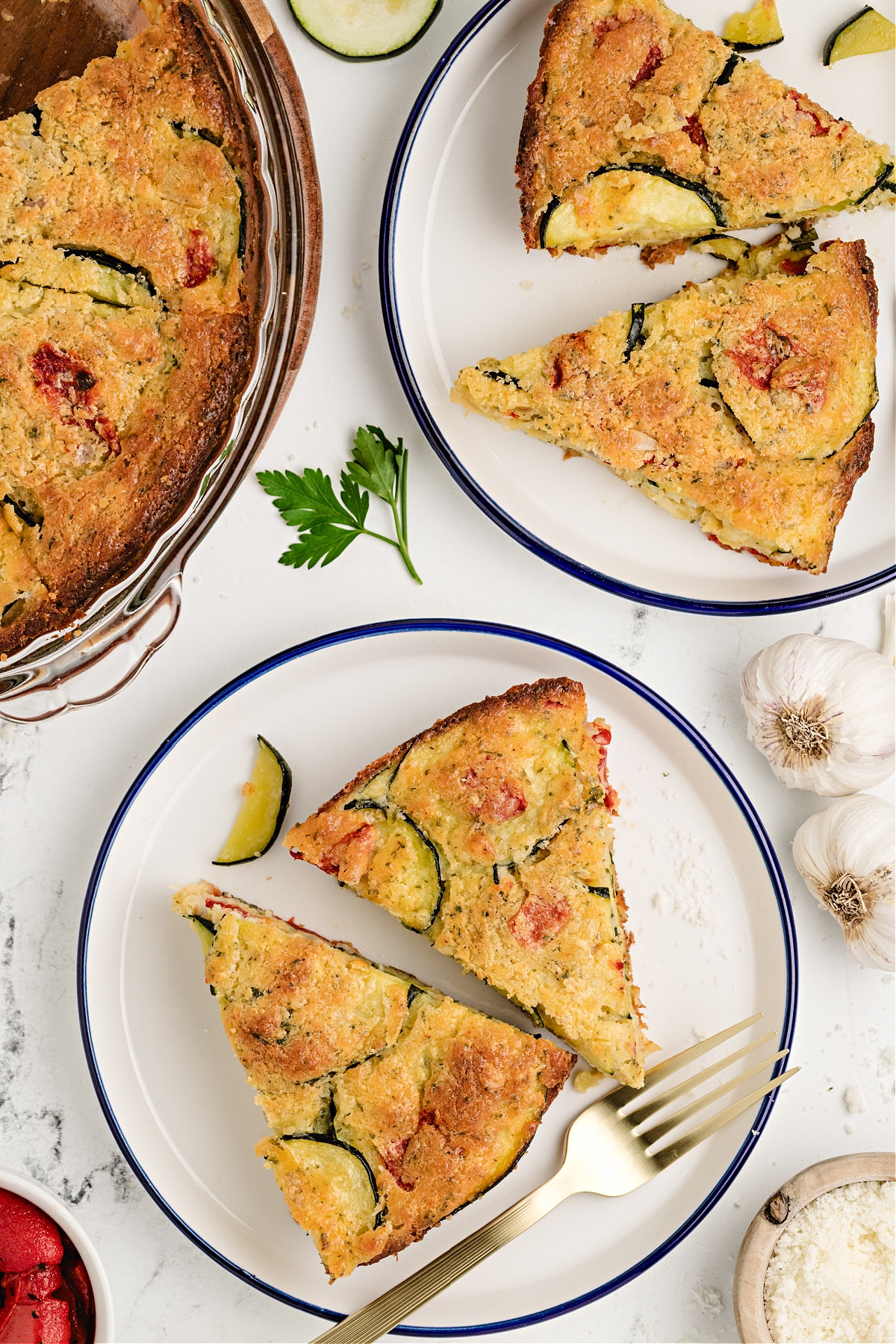 Slices of savory zucchini pie on white plates with garlic, parsley, and roasted pepper on the side