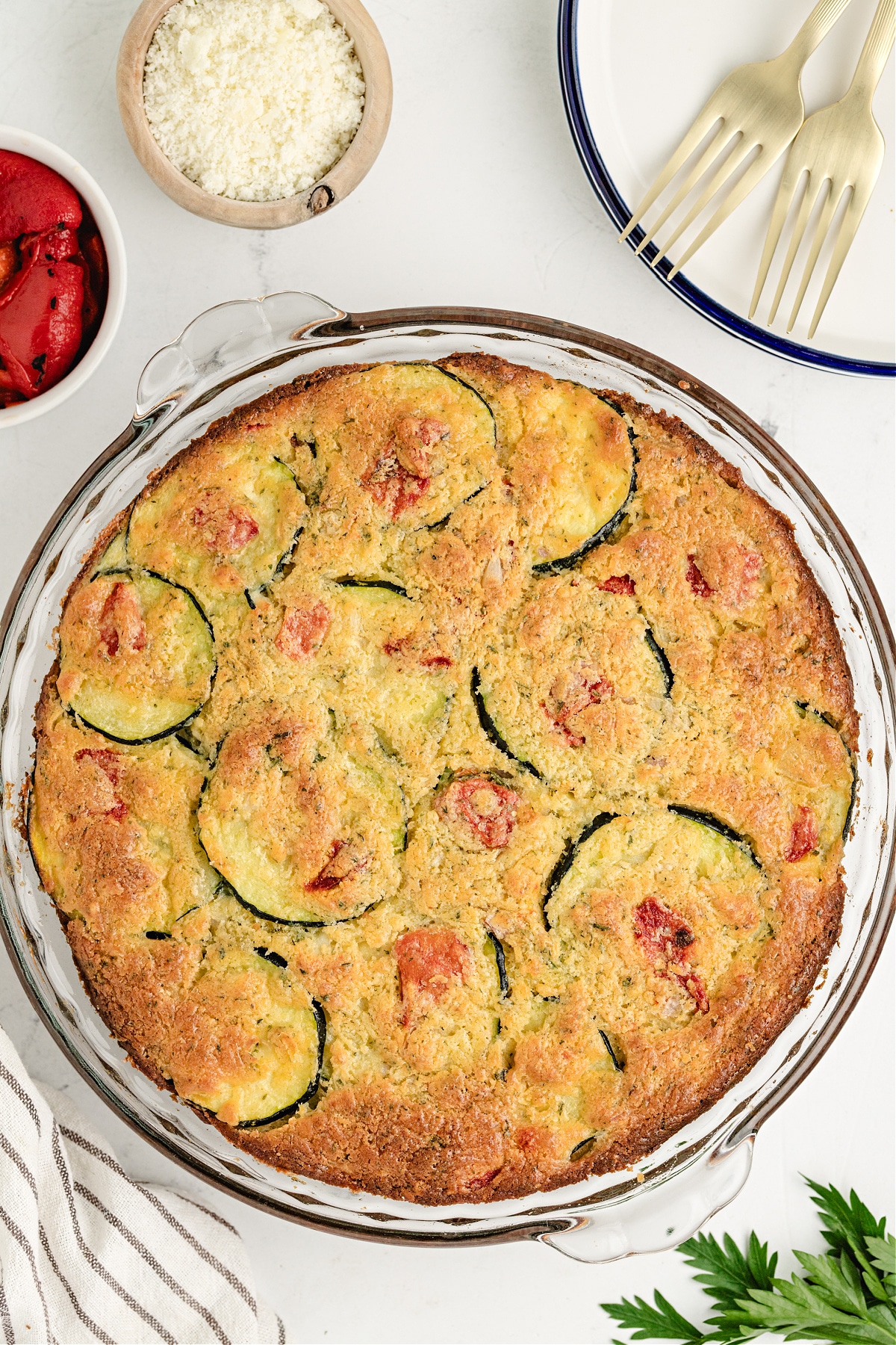 Overhead shot of baked zucchini cheese pie ready to slice, with bowls of roasted pepper and grated cheese