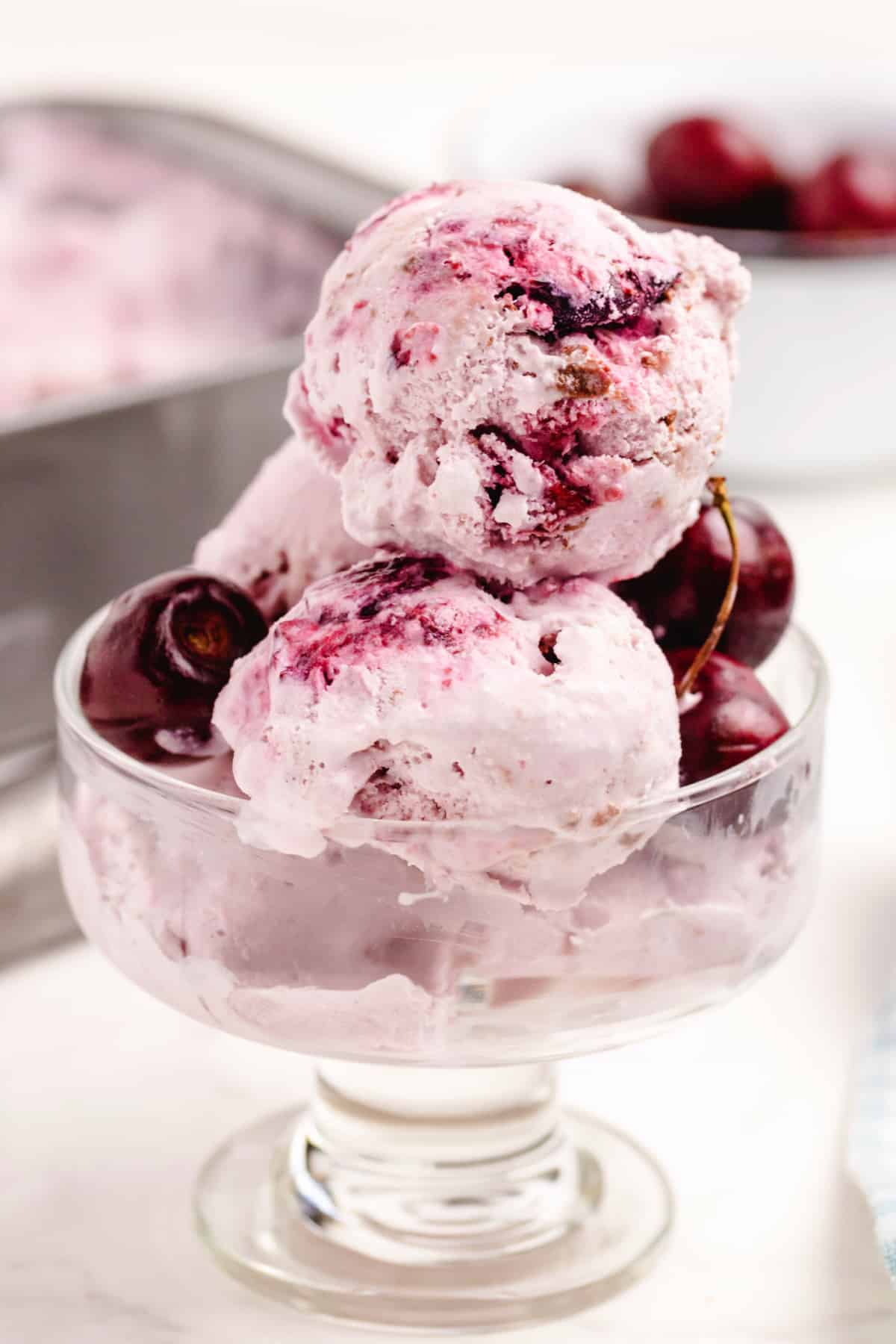 Cherry Coconut Chocolate Chip Ice Cream in Dish Glass dish with scoops of homemade cherry coconut chocolate chip ice cream, showing swirls of cherry and chunks of chocolate.