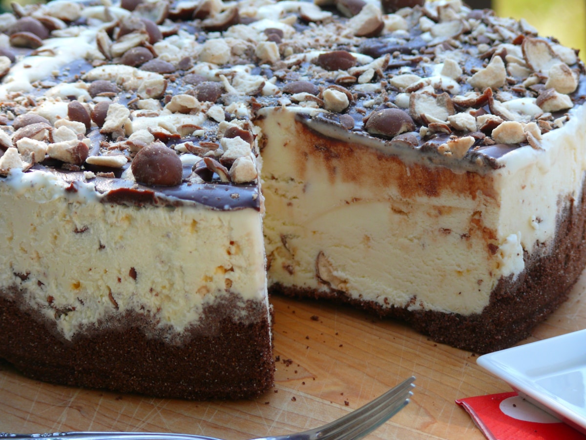 Whoppers Malted Milk Ice Cream Cake layered with vanilla malt ice cream and crushed Whoppers, topped with chocolate ganache and more malted milk balls. Whoppers Malted Milk Ice Cream Cake in a springform pan with a slice removed, showing the vanilla malt ice cream filling, chocolate ganache topping, and chocolate graham cracker crust.