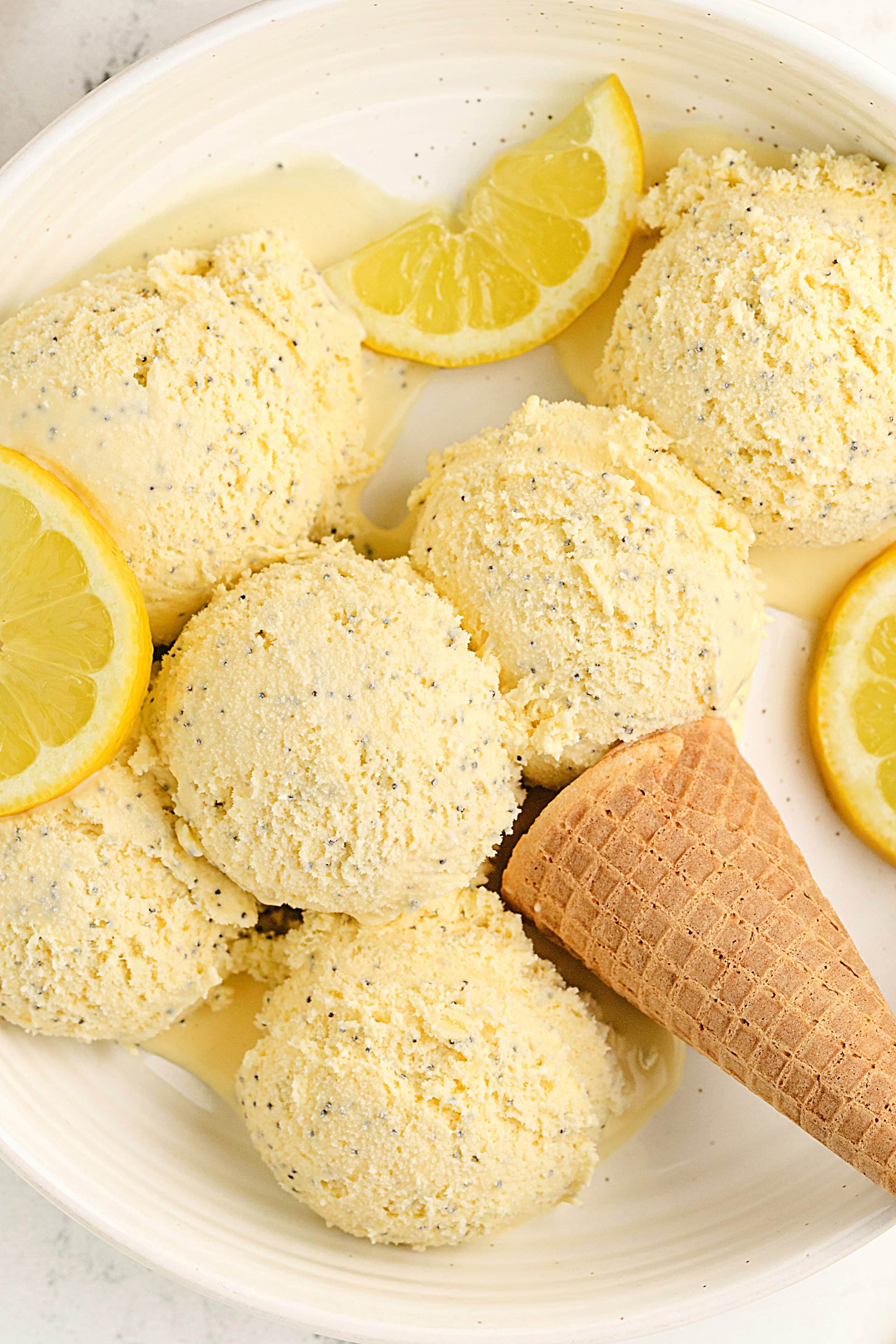 Lemon Curd Ice Cream with Poppy Seeds in a Serving Bowl Scoops of lemon poppy seed ice cream with lemon slices and a cone in a shallow white bowl