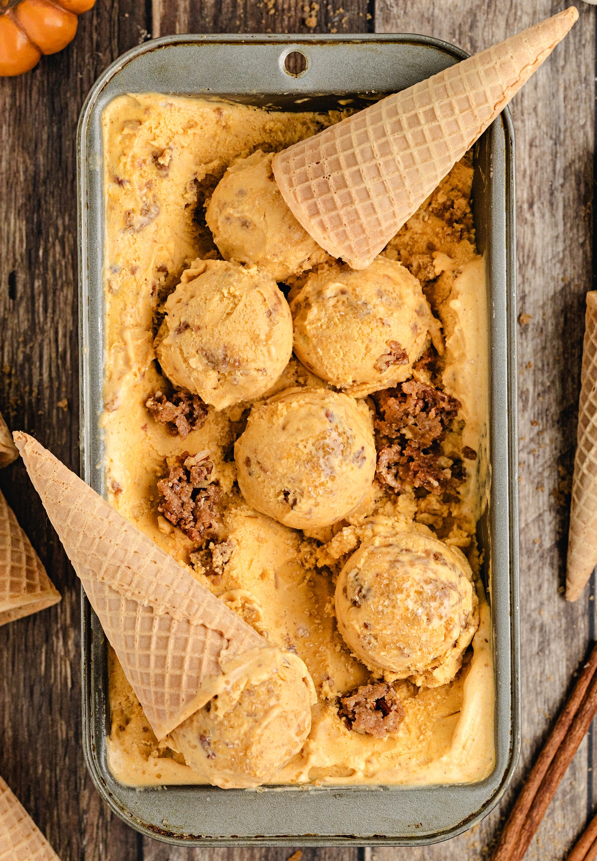 Pumpkin Brown Sugar Crunch Ice Cream with Waffle Cones Scoops of Pumpkin Brown Sugar Crunch Ice Cream in a metal pan with waffle cones and baked streusel pieces, showing creamy texture and rich brown sugar color.