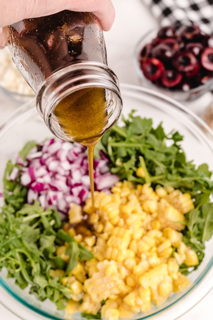 pouring balsamic vinaigrette on cherry corn salad Vinaigrette being poured over arugula, corn, and red onion in a glass mixing bowl.
