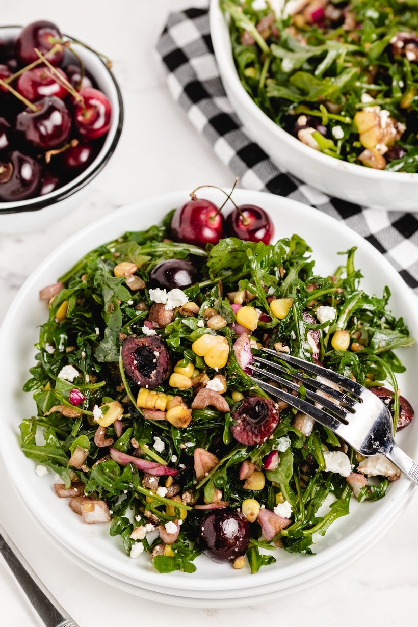 plated cherry corn salad with arugula and feta Plate of cherry corn salad with arugula, feta, and fresh cherries, served with a fork and extra cherries on the side.