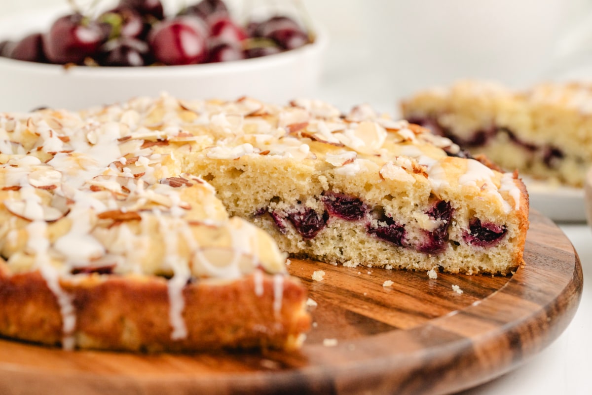 Fresh Cherry Almond Coffee Cake with Glaze and Almond Topping Whole cherry almond coffee cake on a wooden board with one slice cut out, showing baked cherries and slivered almonds
