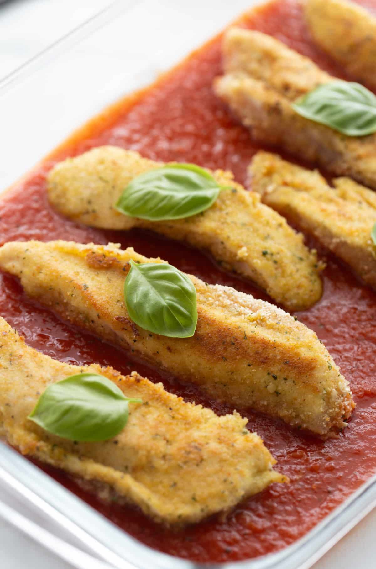 Chicken tenderloins layered in sauce with basil Pan-fried chicken tenderloins placed in marinara with basil leaves on top