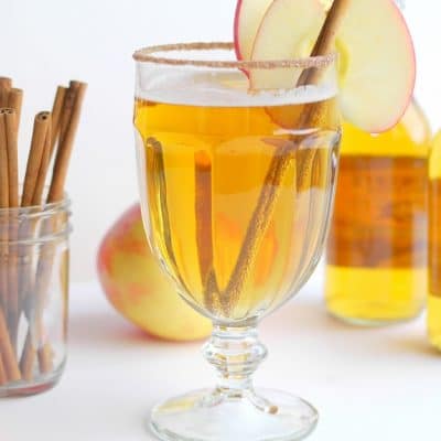 Beer cider slam in a tall glass with a cinnamon stick and slice of apple. Bottle of cider in the background along with cinnamon sticks in a jar.