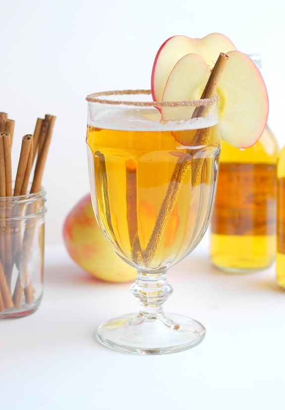 Beer cider slam in a tall glass with a cinnamon stick and slice of apple. Bottle of cider in the background along with cinnamon sticks in a jar.