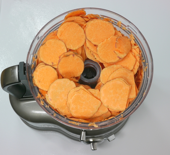 Sliced sweet potatoes sitting in the bowl of a food processor.