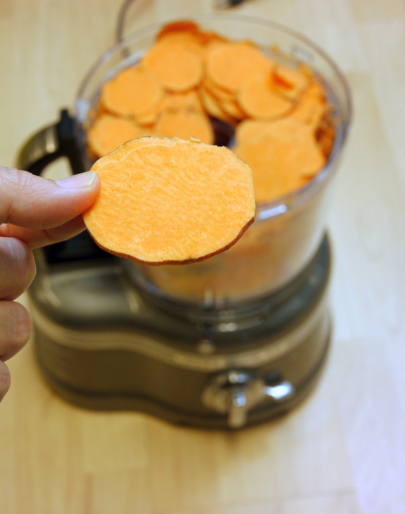 Sliced sweet potato being held by a hand with more in a food processor in the background.