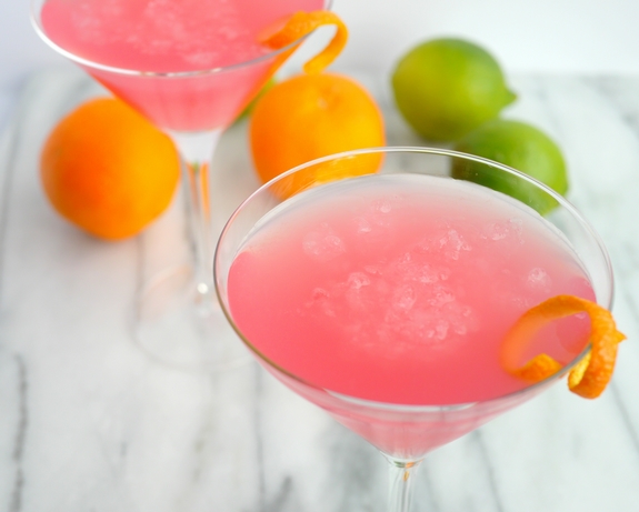 Why would you want to drink anything other than the World's Best Cosmopolitan Cocktail? The only thing girly about this cosmo drink is its color. World's Best Cosmopolitan Cocktail in two martini glasses with limes and oranges in the background.