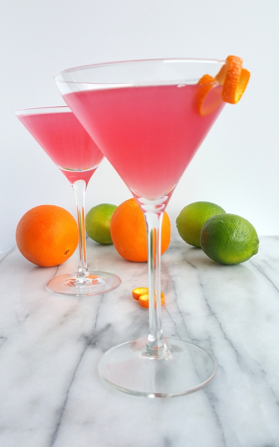 Why would you want to drink anything other than the World's Best Cosmopolitan Cocktail? The only thing girly about this cosmo drink is its color. World's Best Cosmopolitan Cocktail in martini glasses with limes and oranges in the background.
