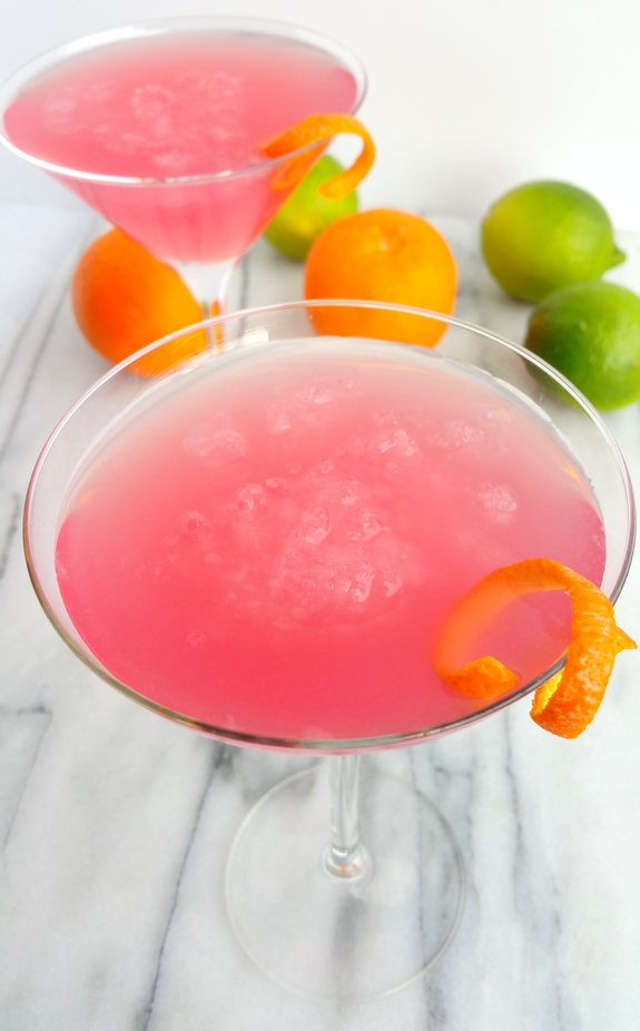 Why would you want to drink anything other than the World's Best Cosmopolitan Cocktail? The only thing girly about this cosmo drink is its color. Two of the World's Best Cosmopolitan Cocktails in martini glasses with limes and oranges in the background.