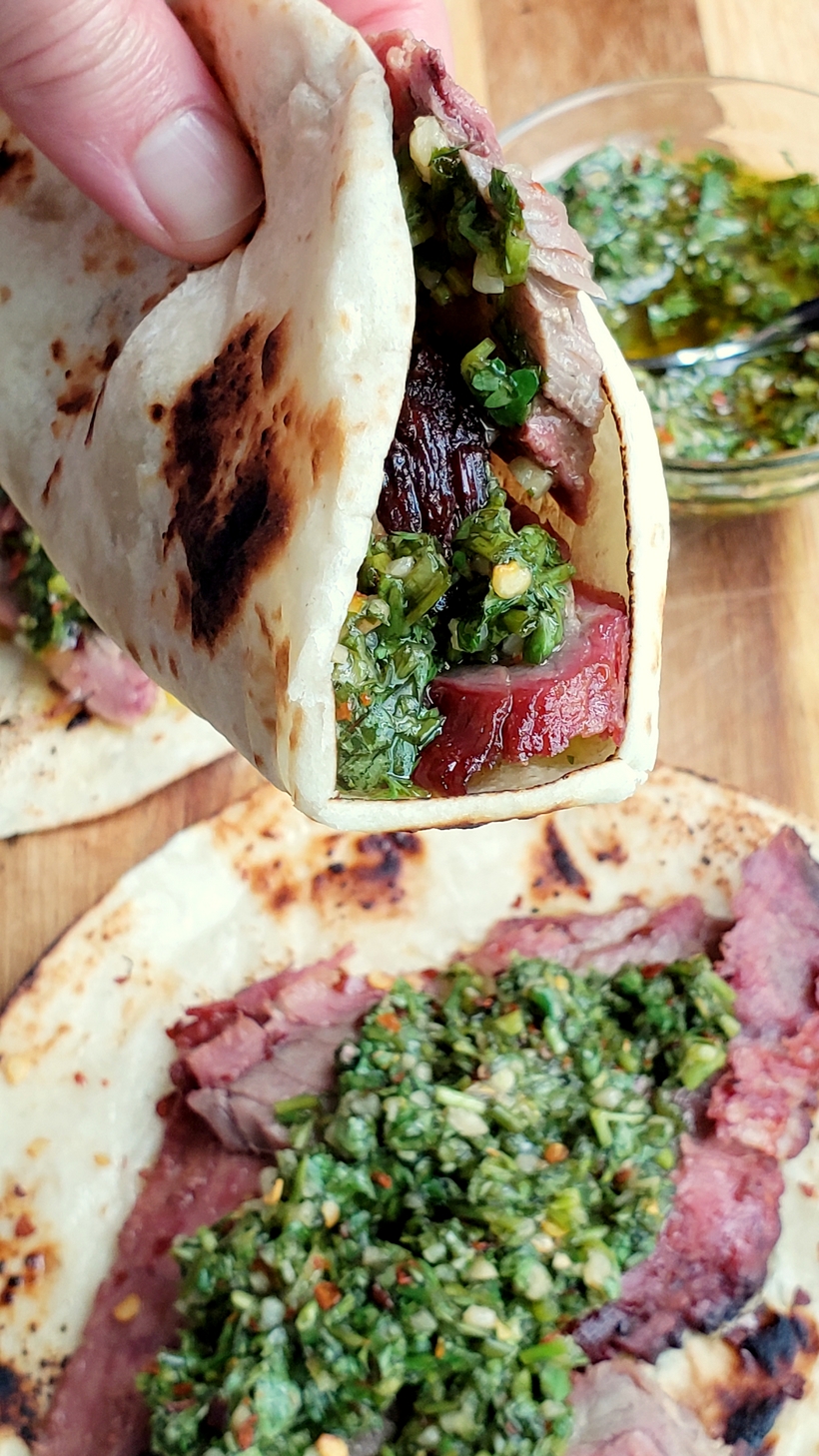 Hand holding a flank steak taco with one below..bowl of chimichurri in the background.
