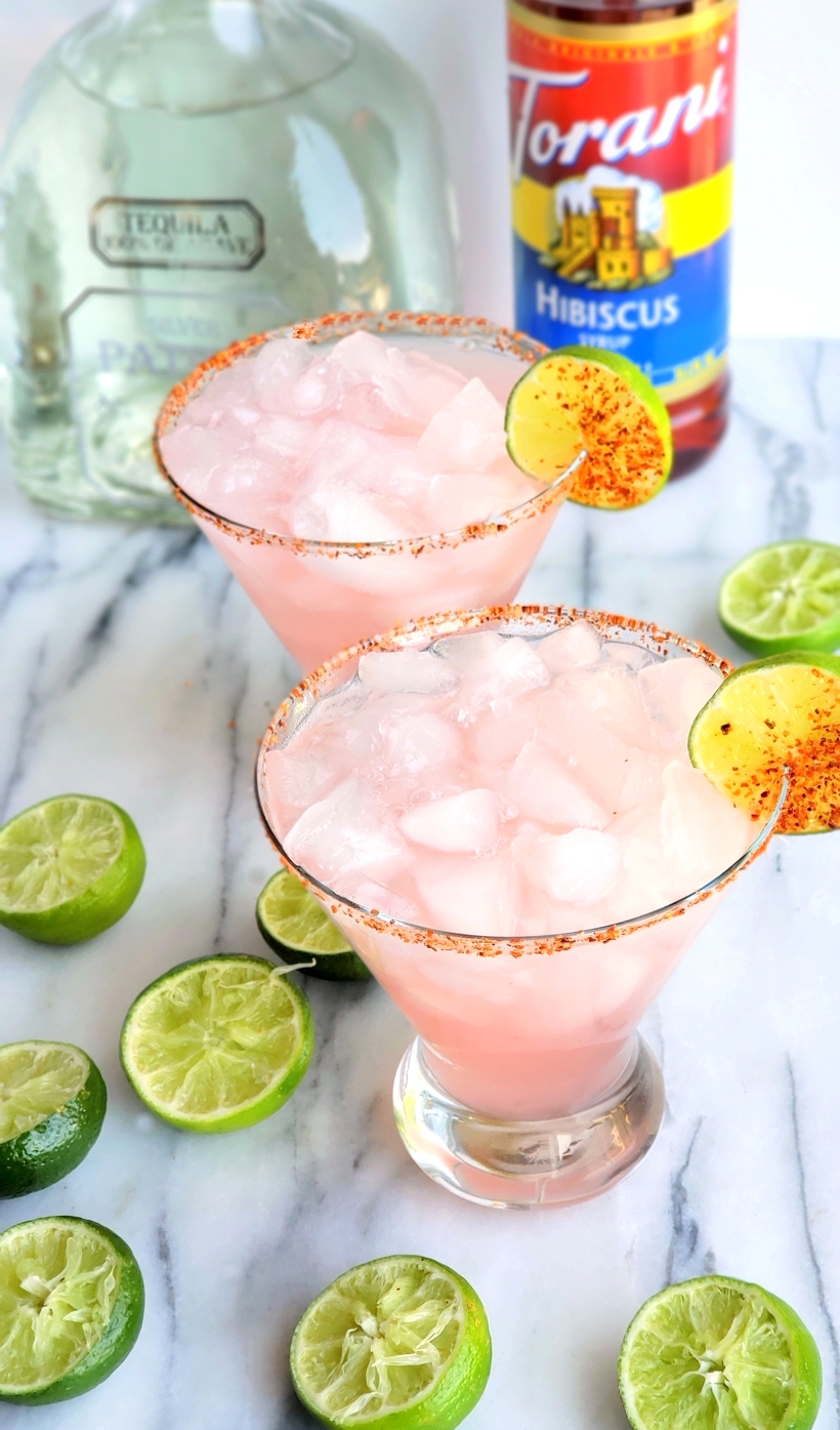 The petal pink color of this Icy #Hibiscus #Margarita is alluring, but it's the sweet, tangy and refreshing flavor that will have you asking for a second pour. #cocktail #noblepig Two hibiscus margaritas with a bottle of torani hibiscus syrup and a bottle of patron tequila with squeezed limes surrounding them.