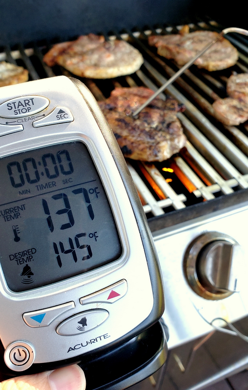 Grilling a quick and easy meal has never been so easy. These tender and Juicy Grilled Pork Chops with Spicy Peach Glaze come together quickly for a dinner your family and friends will not soon forget. The glaze can be put together in minutes, while the pork chops undergo a quick brine. #noblepig #pork #porkchops #grilling #easydinners Pork chops cooking on a grill with a digital meat thermometer taking their temperature.