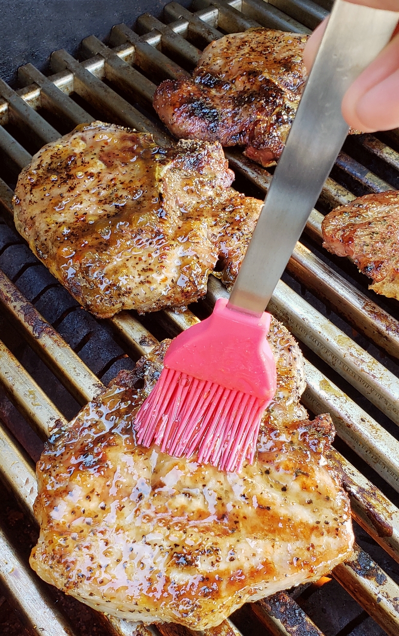 Grilling a quick and easy meal has never been so easy. These tender and Juicy Grilled Pork Chops with Spicy Peach Glaze come together quickly for a dinner your family and friends will not soon forget. The glaze can be put together in minutes, while the pork chops undergo a quick brine. #noblepig #pork #porkchops #grilling #easydinners Pork chops on a grill being basted with a brush.