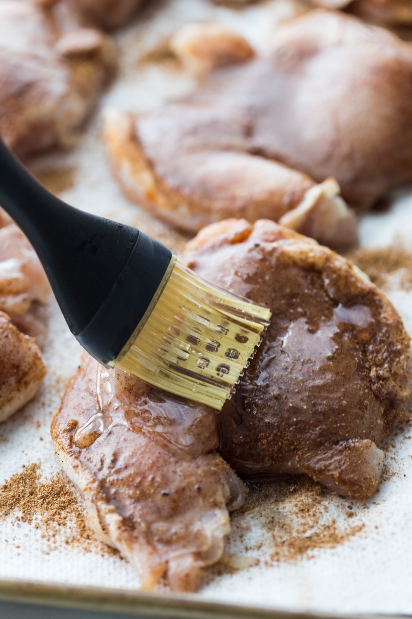 A pastry brush applying melted coconut oil to the spiced chicken thighs before air frying.