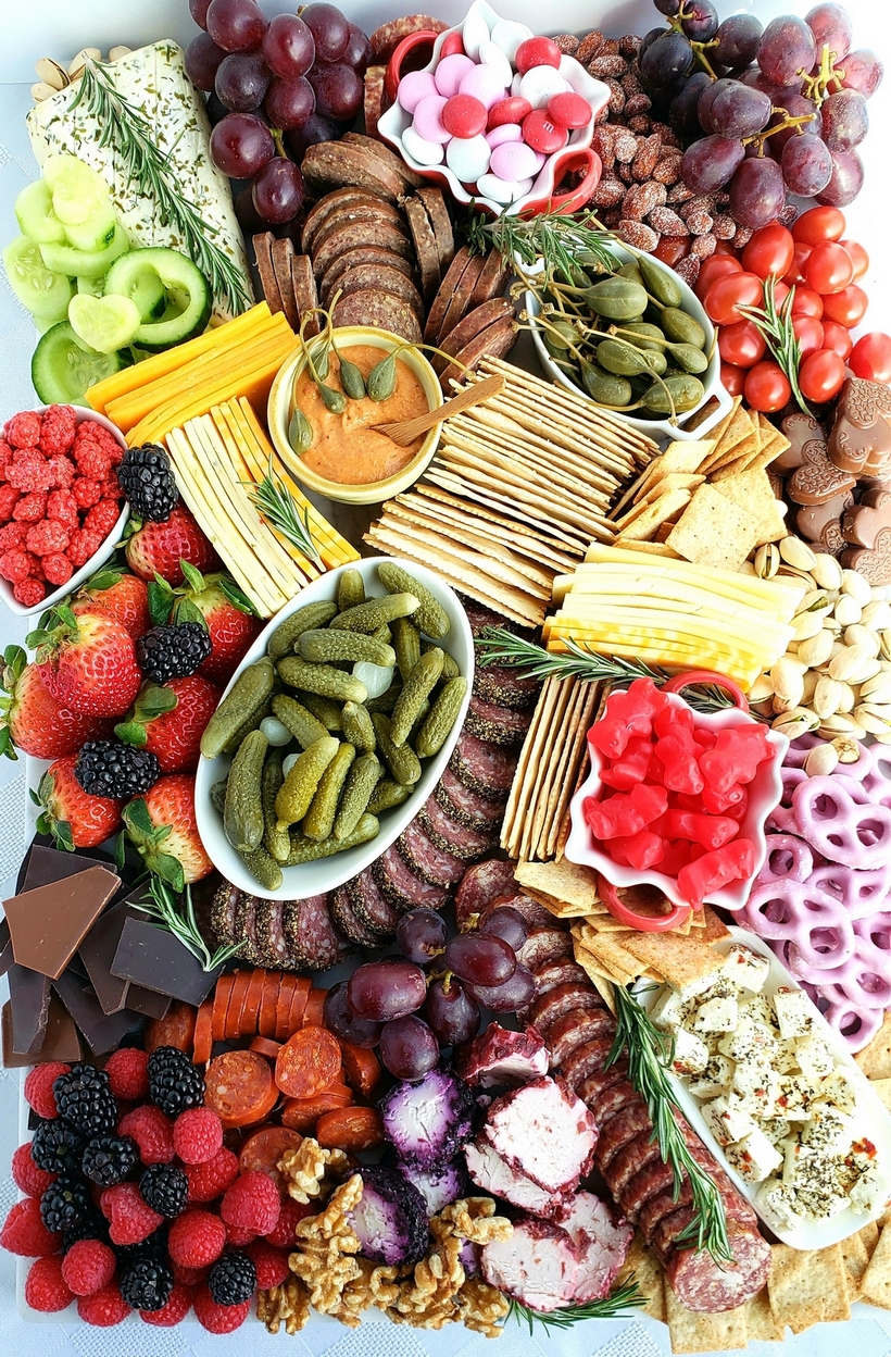 Valentine's Day Charcuterie Board for date night. Overhead view of a Valentine’s Day charcuterie board filled with sliced meats, cheeses, fresh fruit, nuts, and festive sweets, arranged on a wooden board with rosemary sprigs for garnish.