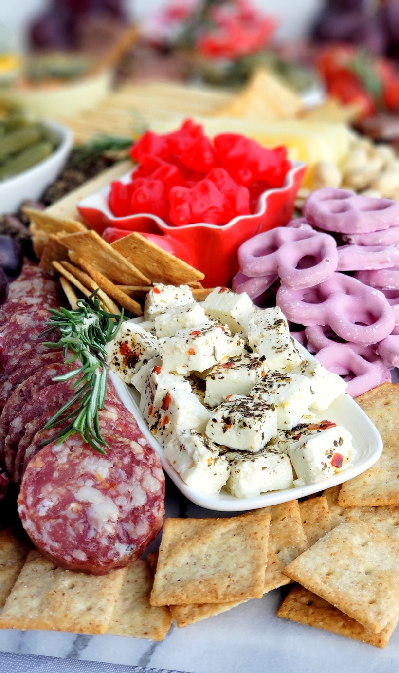 Valentine's Day Charcuterie Board with marinated cheese and pretzels. Close-up of heart-shaped cucumber slices alongside cheddar, Colby, Swiss, and pepper jack cheese, with pepperoni and soppressata fanned out nearby.