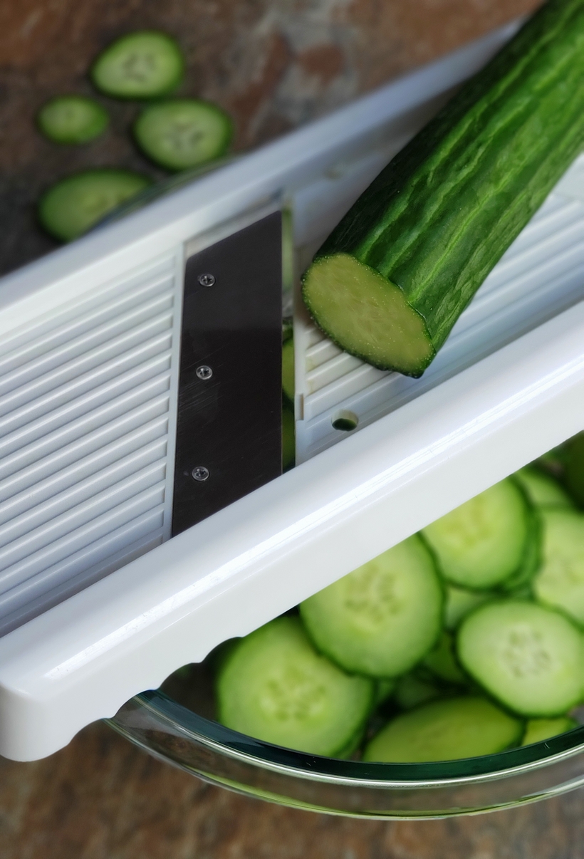 Keep it light and refreshing ~ Cucumbers with Mint Vinaigrette are the perfect side dish for any meal. They are especially wonderful with Asian Cuisine. #cucumbers #sidedish #asiancuisine A cucumber being sliced on a mandoline.
