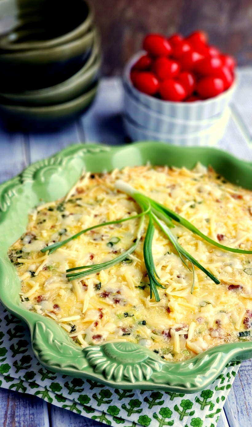 A decadent and sumptuous brunch recipe, this Mouthwatering Oven Omelet will have you wondering why you make eggs any other way. Everyone at your breakfast table will imagine you fussed over this easy omelet, when in reality this egg dish takes very little time to put together. #noblepig #ovenomeletterecipe #ovenomelet #breakfastcasserole #brunchrecipe #bakedomelette #bakedomelet #bruncheggs #bruncheggsforacrowd #bruncheggideas #breakfastfordinner #bruncheggscasserole #brunchideas Oven Omelet on a printed napkin in a green dish with tomatoes and empty bowls in the background.