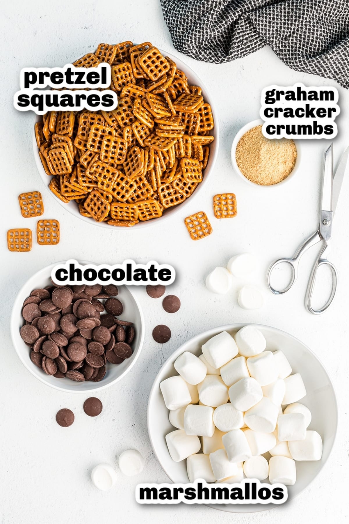 Ingredients for s’mores pretzels including square pretzels, chocolate melting wafers, graham cracker crumbs, and marshmallows arranged in bowls