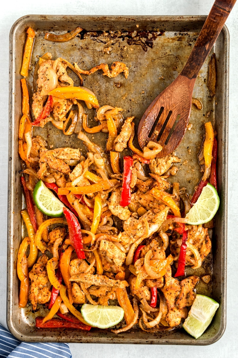 A wooden spoon scooping out Sheet Pan Chicken Fajitas A sheet pan of fajitas with some of the chicken and vegetables scooped up by a wooden spoon, showing the texture and color of the dish.
