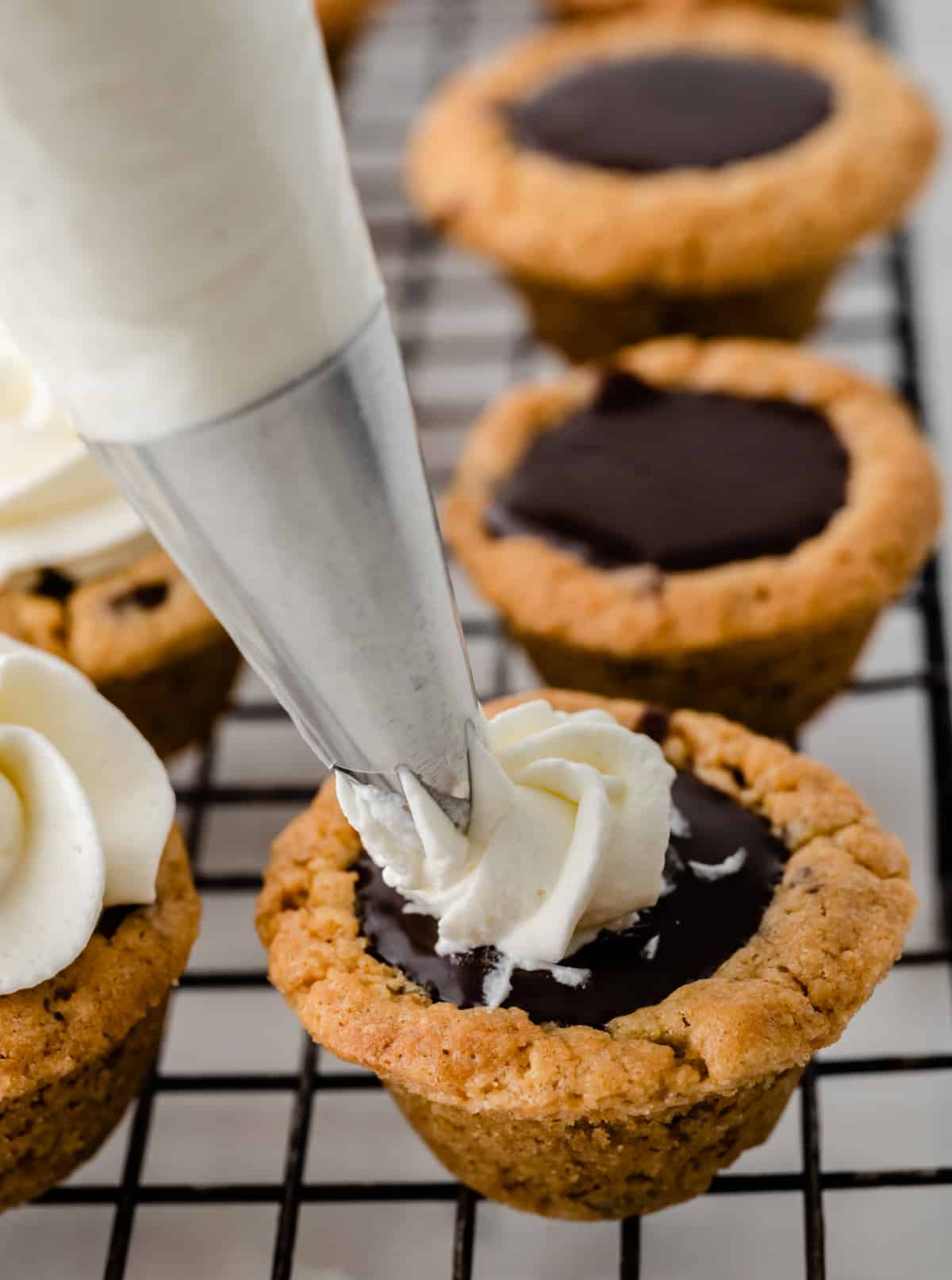 Nutella chocolate cookie cups being topped with whipped cream on a cooling rack