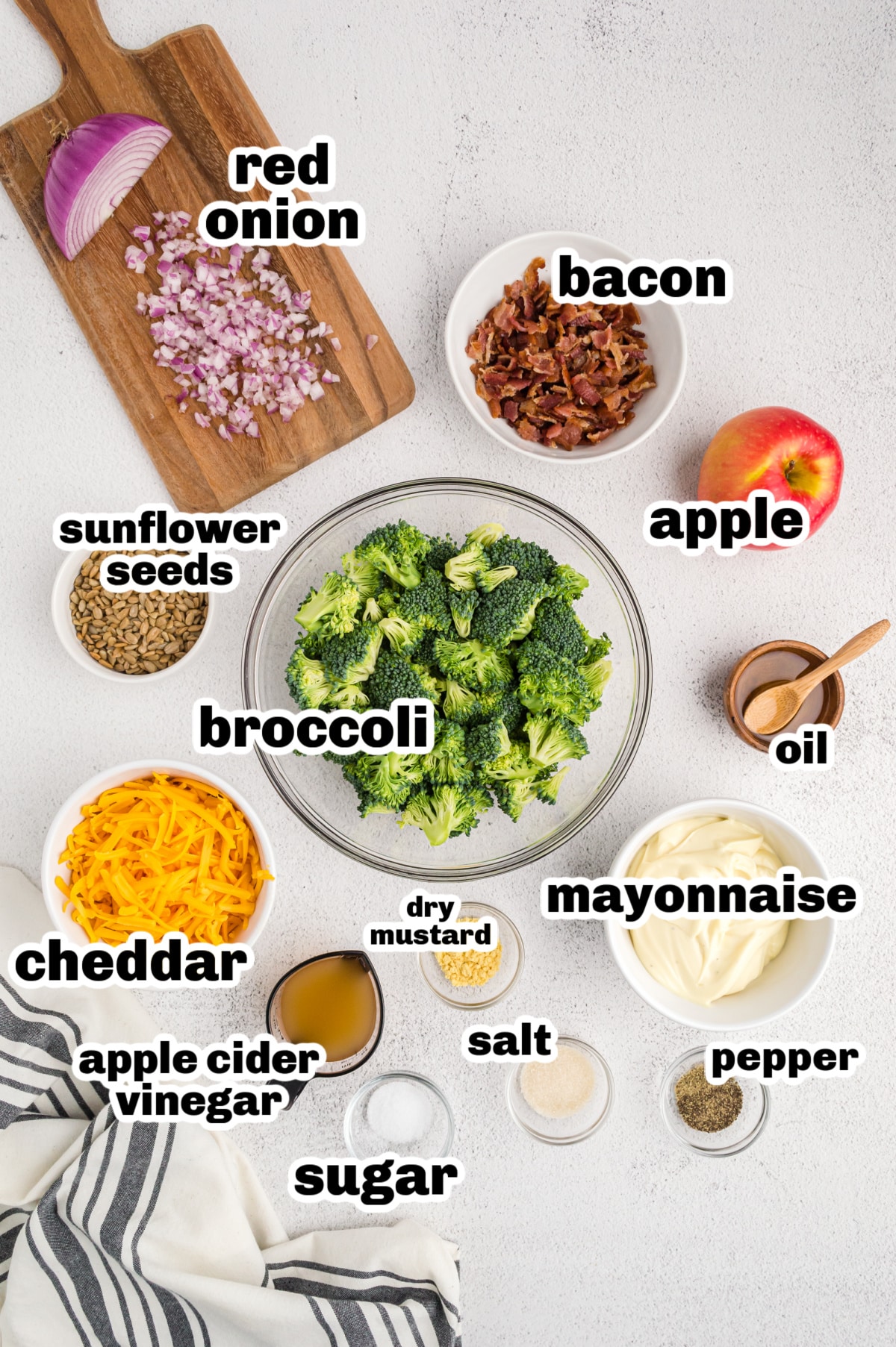 Bacon Broccoli Salad ingredients A top-down view of all the ingredients for bacon broccoli salad arranged on a countertop, including chopped broccoli, bacon, cheese, apple, sunflower seeds, and dressing ingredients.