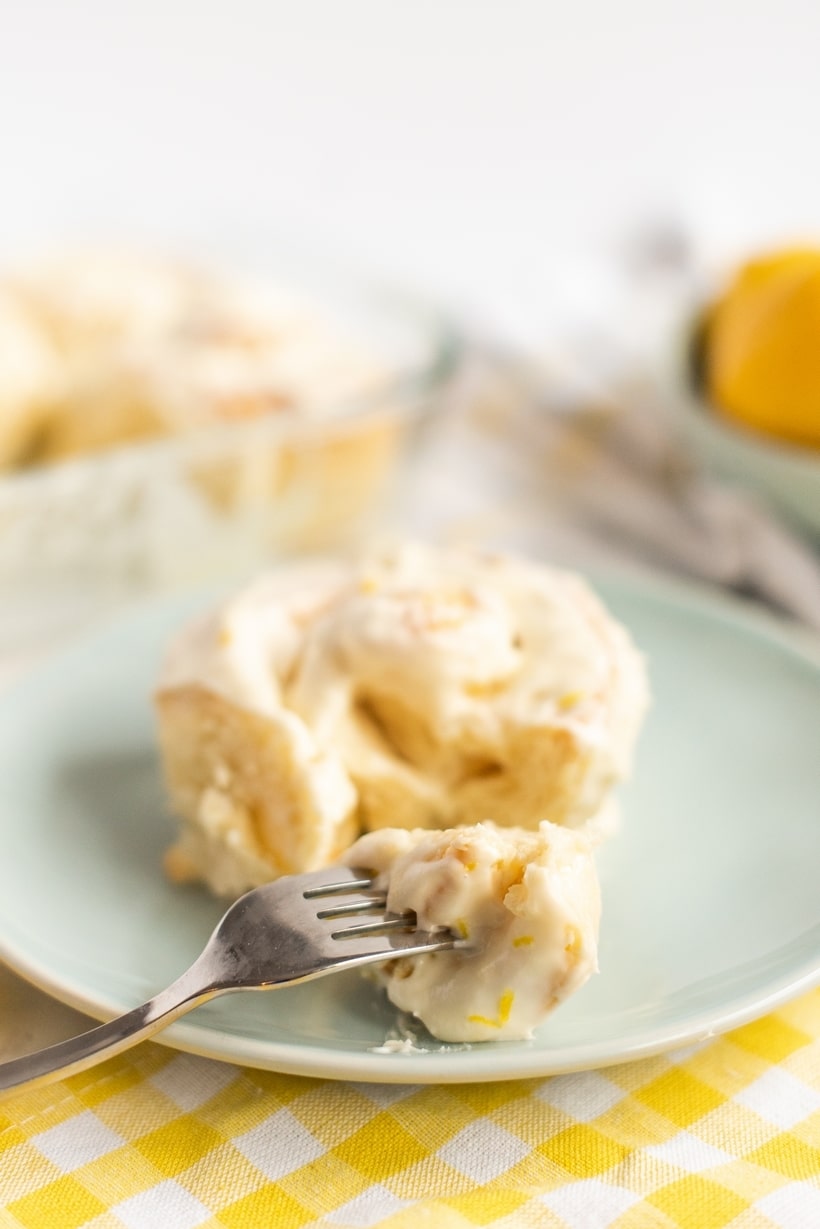 Lemon Buns with Lemon Cream Cheese Icing on a plate. A soft, fluffy lemon bun with cream cheese icing on a white plate, with a fork lifting a bite-sized piece.