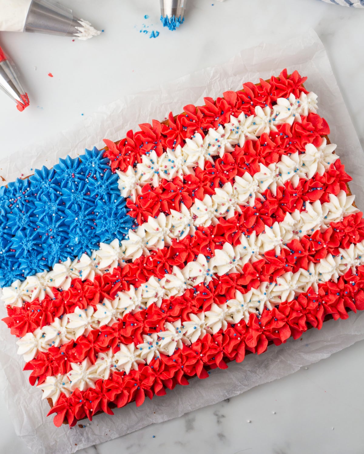  Overhead view of buttercream American flag cake decorated with piped red, white, and blue stars, piping bags visible in the background.