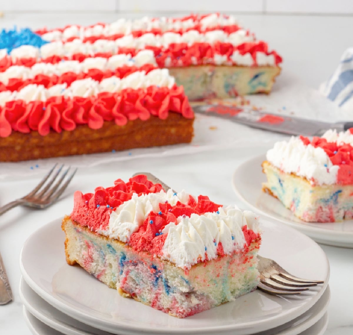 Two slices of patriotic flag cake with piped red, white, and blue frosting and swirl pattern inside, full cake in background.