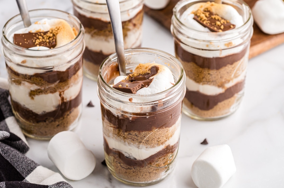 Multiple no-bake s’mores dessert jars on a white surface with chocolate, graham cracker, and marshmallow layers