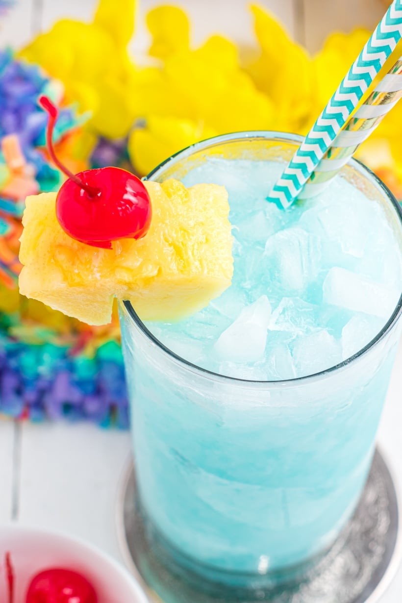 This Blue Hawaiian cocktail tastes like a vacation in a glass. Notes of pineapple, coconut and citrus from the Blue Curacao liqueur make it the perfect tropical cocktail for summer parties! close-up of a blue curacao cocktail with crushed ice and a pineapple and cherry wedge with a straw
