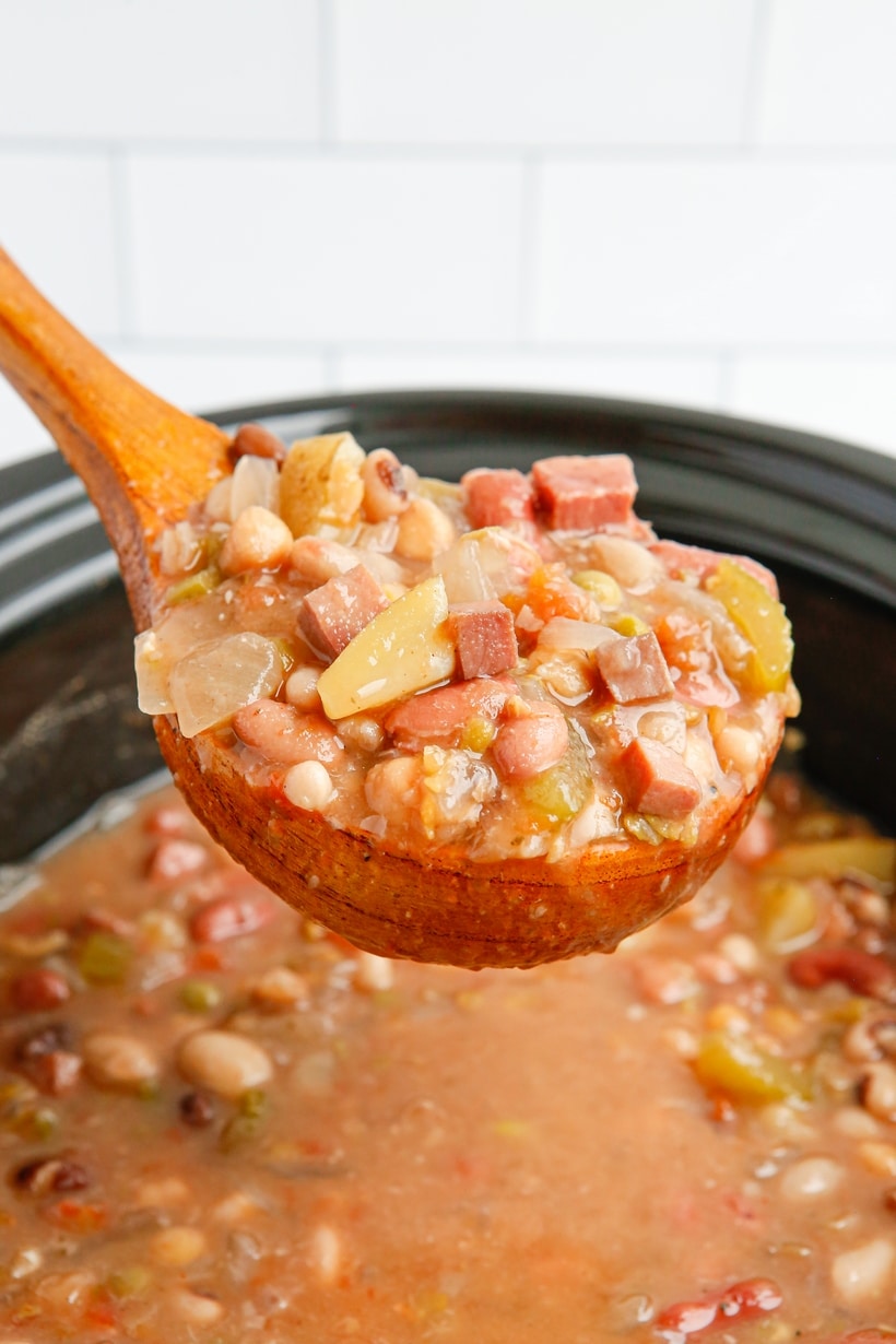 A wooden ladle lifting a hearty scoop of ham and bean soup from the slow cooker.