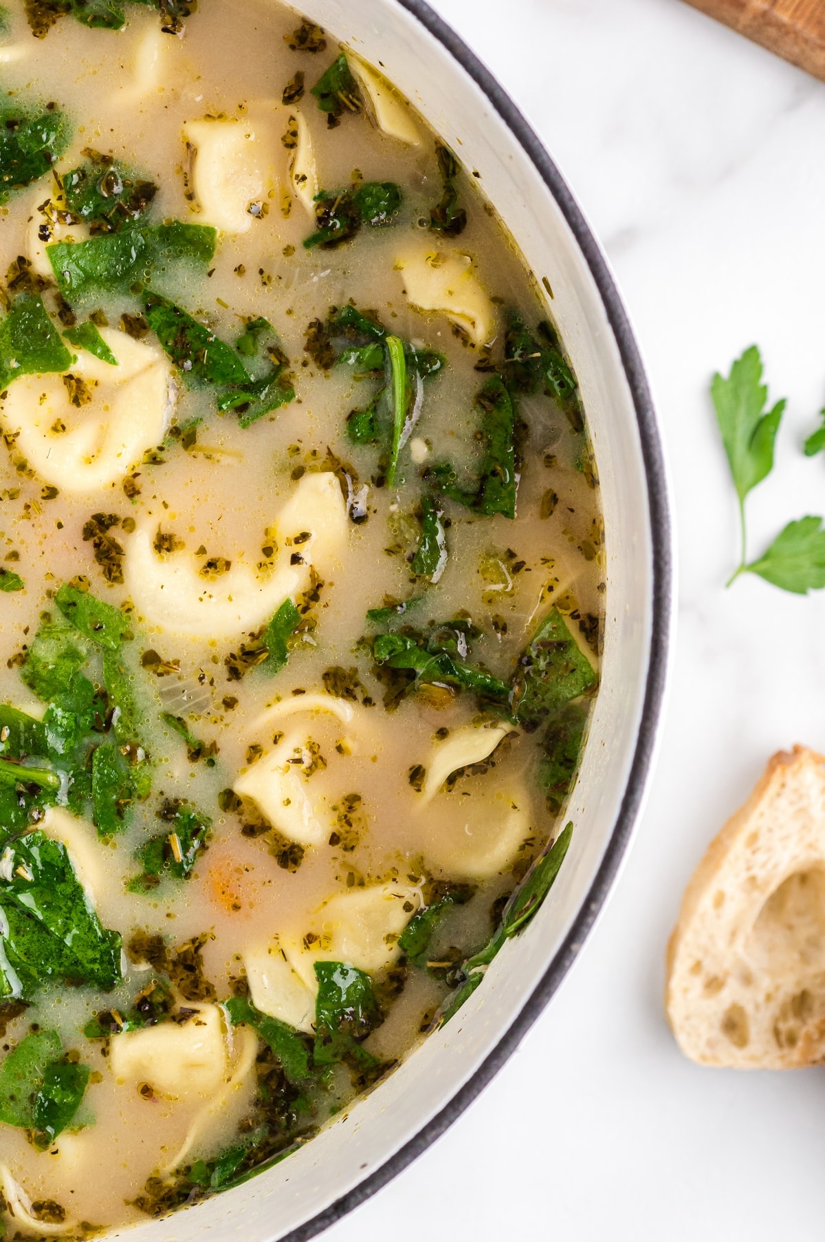 A close-up view of soup in a white pot showing cheese tortellini, spinach, herbs, and broth along the rim of the pot.